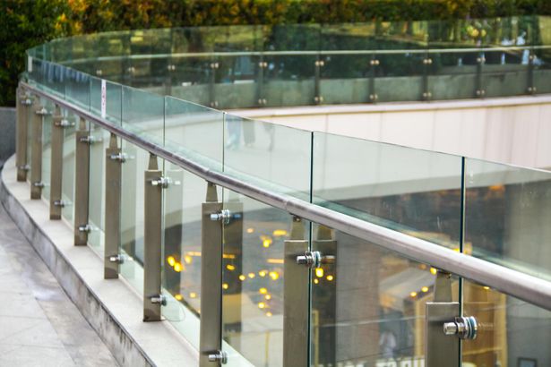 Glasgeländer mit Metallhandlauf auf Betonboden. Das Geländer geht rechts in einer Rundung auf einer höheren Ebene eines Gebäudes entlang 