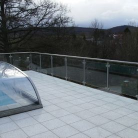 große Terrasse mit Glasgeländer mit Blick auf eine grüne Fläche mit Baum und Häusern im Hintergrund.