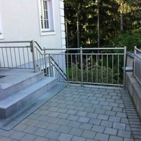 klassisches Metallgeländer an einer Treppe mit zwei Stufen, die auf einen mit Steinen gepflasterten Weg mit Handgeländer führt. Im Hintergrund ist ein Teil eines Hauses mit Fenster zu sehen.