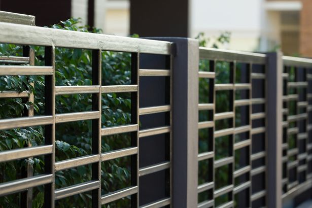 modernen Zum aus Metall rechteckiges Design. Die Zaunstreben bestehen aus horizontalen Rechtecken in verschiedener Höhe. Hinter dem Zaun ist eine Hecke zu sehen.